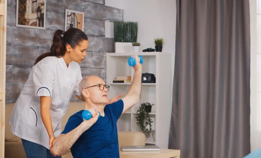 A therapist working through a seated dumbbell exercise with a patient at home